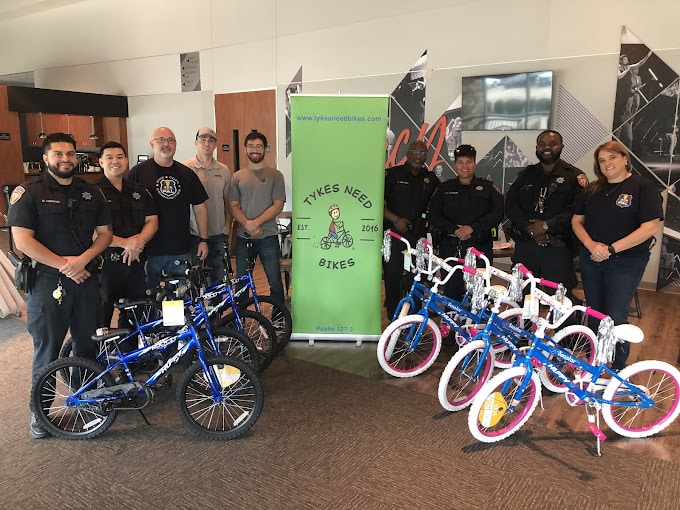 Group of police officers and community members standing next to a green “Tykes Need Bikes” banner and several new children’s bicycles, including blue and pink bikes, in an indoor setting.