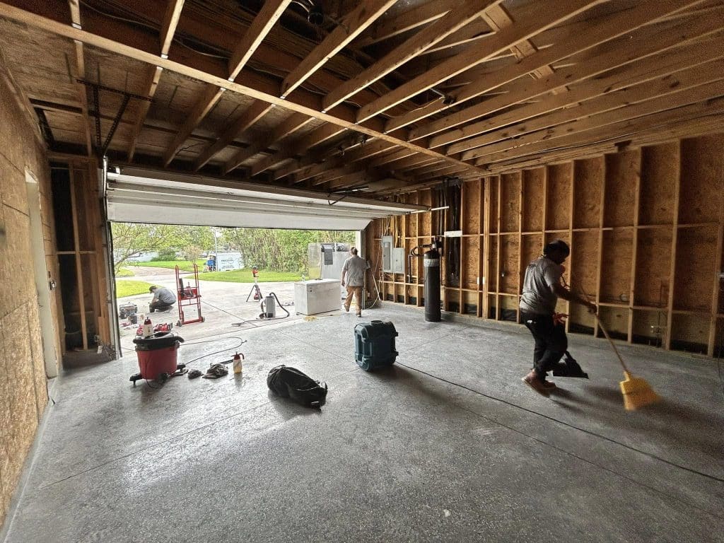 A garage with exposed wooden framing and a wet concrete floor is in the process of water damage restoration in Houston, TX. Workers are sweeping, and drying equipment is placed throughout the space. The garage door is open, showing an outdoor view. Type residential water damage restoration near me in Houston, TX