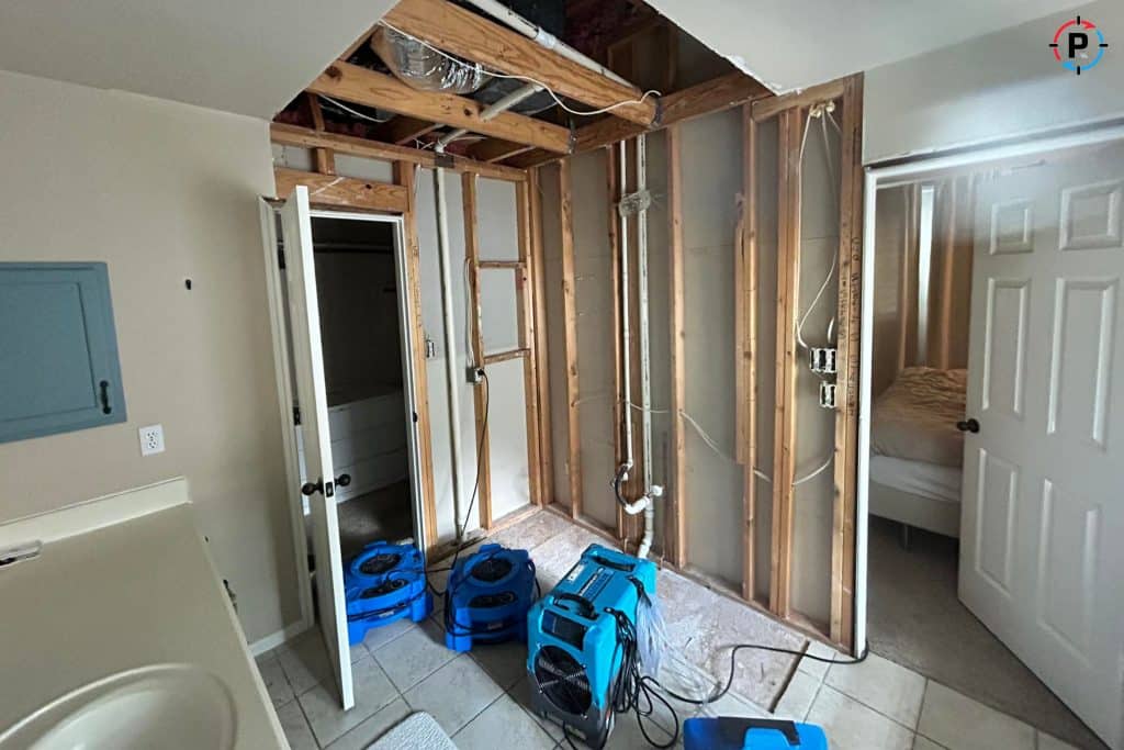 A residential property room undergoing water damage restoration in Houston. The drywall has been removed, exposing wooden framing and plumbing. Air movers and a dehumidifier are positioned to dry out the affected area. The tile flooring appears intact, and a partially open door leads to another room.