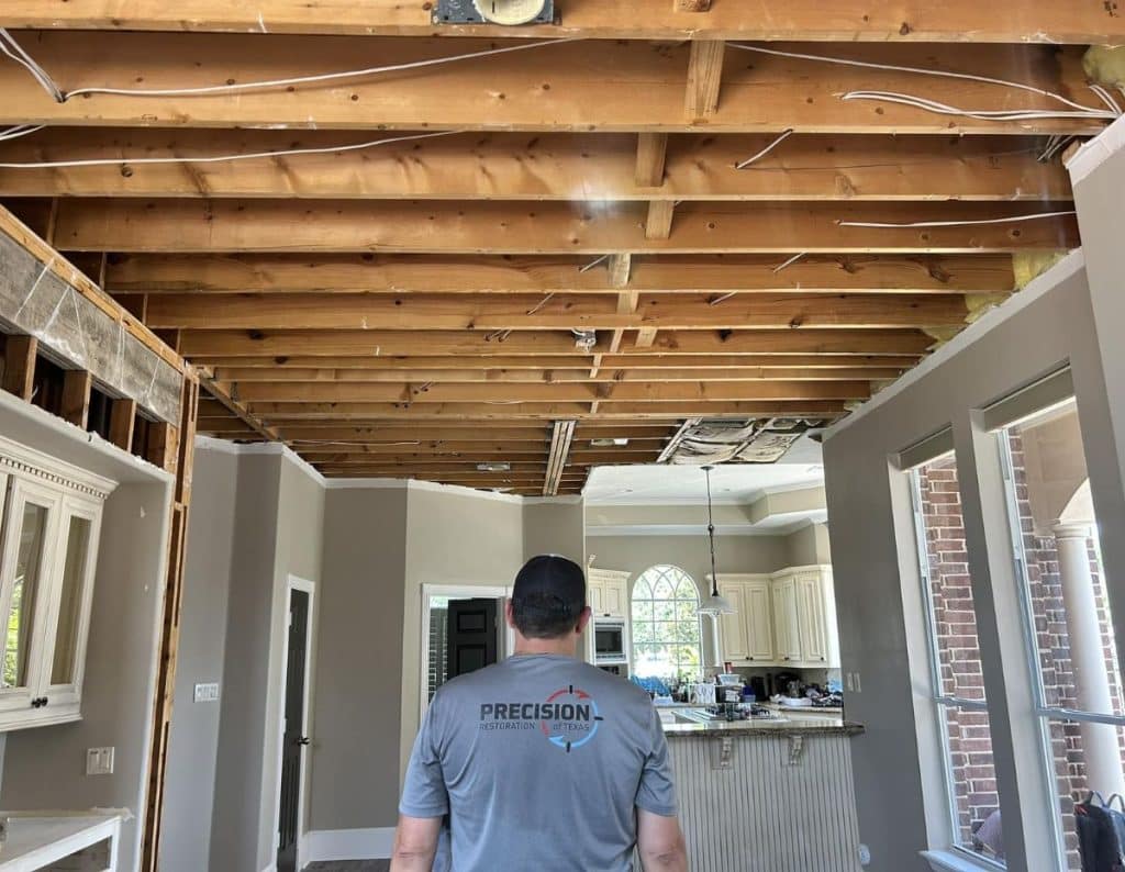 What do a restoration company do? A restoration professional from Precision Restoration Texas assesses water damage in a residential kitchen, with exposed ceiling beams and visible structural repairs in progress in Houston, TX