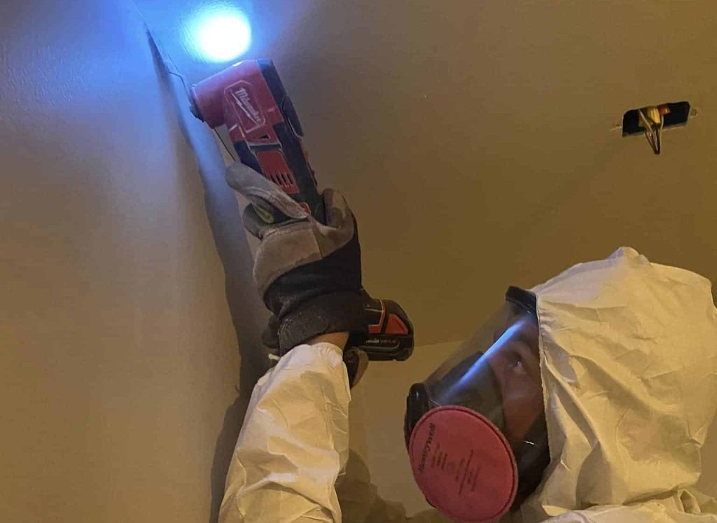 A mold inspection professional wearing a white hazmat suit and respirator is using a thermal imaging camera to check for moisture issues on a wall. The image is part of a promotional card with a red background that highlights 'INSPECTION AND ASSESSMENT.' The text explains how thermal imaging cameras, moisture meters, and licensed mold assessment consultants help identify moisture sources and mold types. This service is essential for mold remediation Houston and mold removal Houston, ensuring thorough detection and assessment of mold-related issues.