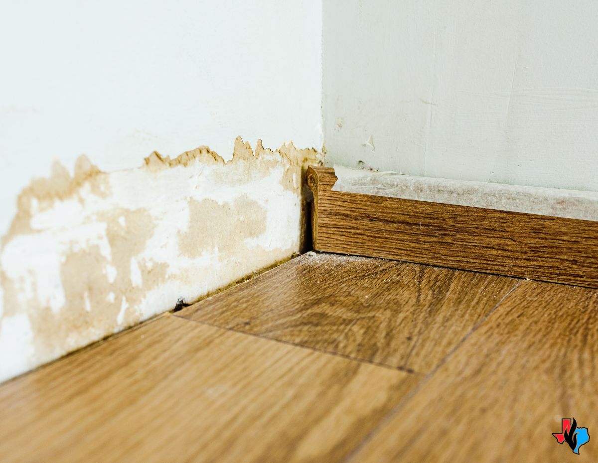 Signs of water damage under floor—wooden floorboards lifting and separating near a damaged wall with peeling paint and discoloration, indicating moisture issues.