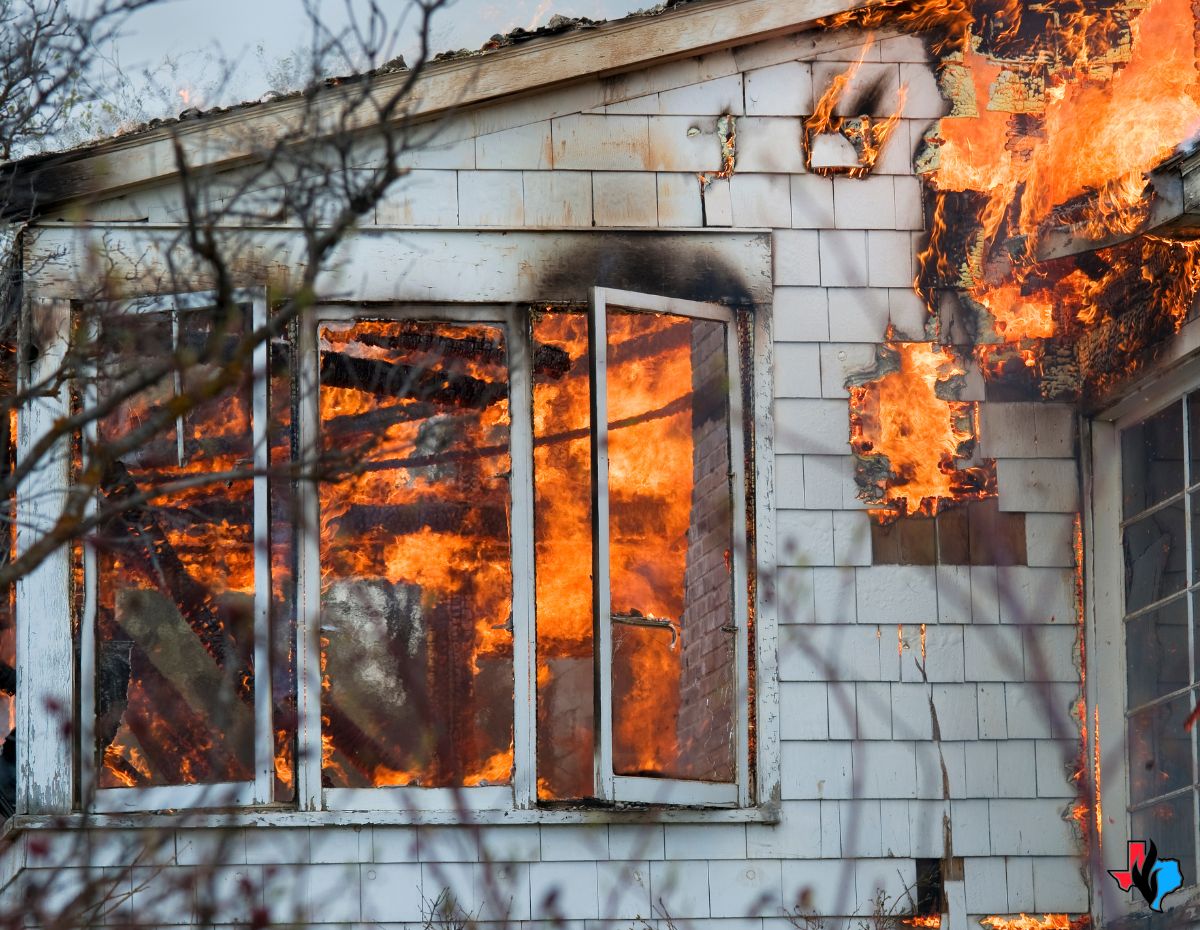 A house fire with intense flames coming out of broken windows and charred siding, symbolizing the aftermath of a disaster. This image highlights the destruction caused by fire and emphasizes the importance of knowing what to do after a kitchen fire to ensure safety and begin the restoration process.