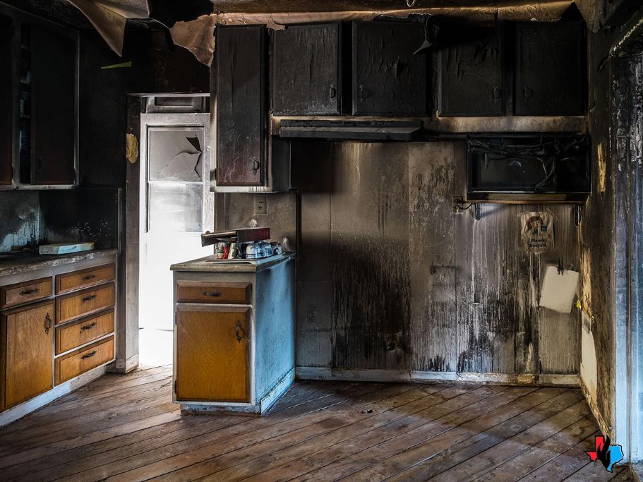 A severely fire-damaged kitchen with blackened cabinets, scorched walls, and charred surfaces, showing the aftermath of an intense house fire.