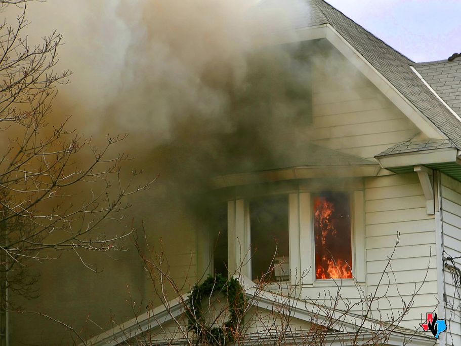 Thick, dark smoke pours from the windows of a two-story house as bright orange flames burn inside a lower-level room. The smoke engulfs the exterior siding and roofline, demonstrating how fire and smoke damage can rapidly spread throughout a structure. Bare tree branches frame the scene, emphasizing the chaos and urgency of the fire event.