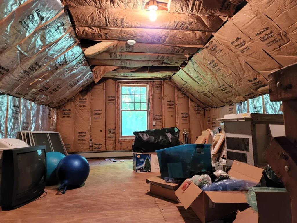 Unfinished room with insulation installed along the sloped ceiling and walls, various stored items on the wooden floor, and a window letting in natural light.
