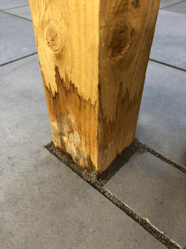 wooden post with significant water damage and rot at its base, where it meets a tiled or paved ground surface.