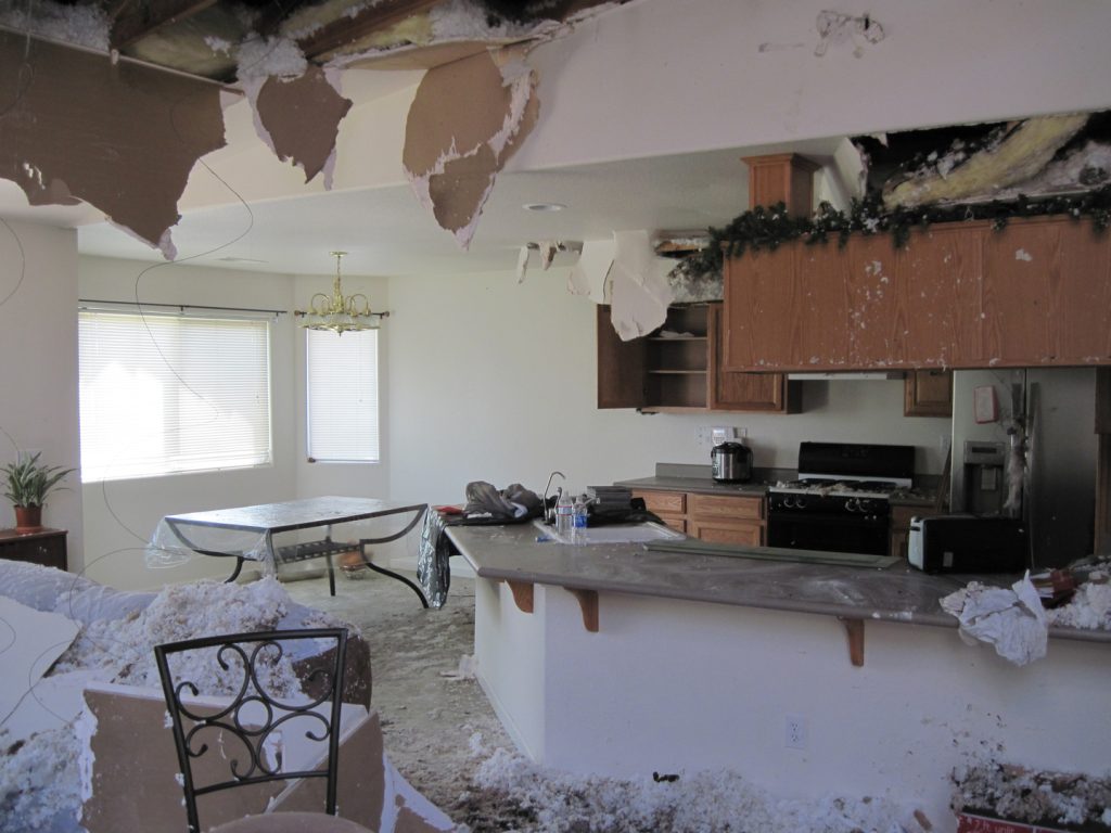 Kitchen and dining area with severe fire and water damage, showing collapsed ceiling, exposed insulation, and debris covering the floor.