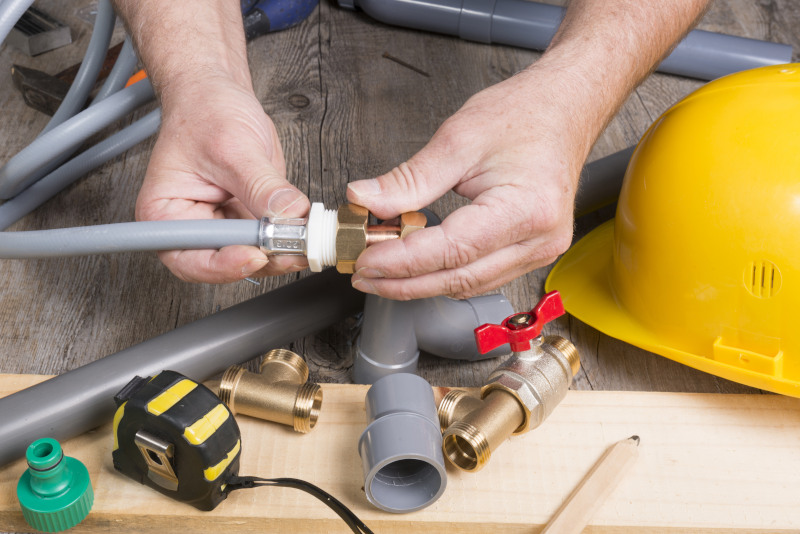 Person working on plumbing pipes with tools and a yellow hard hat on a wooden surface.